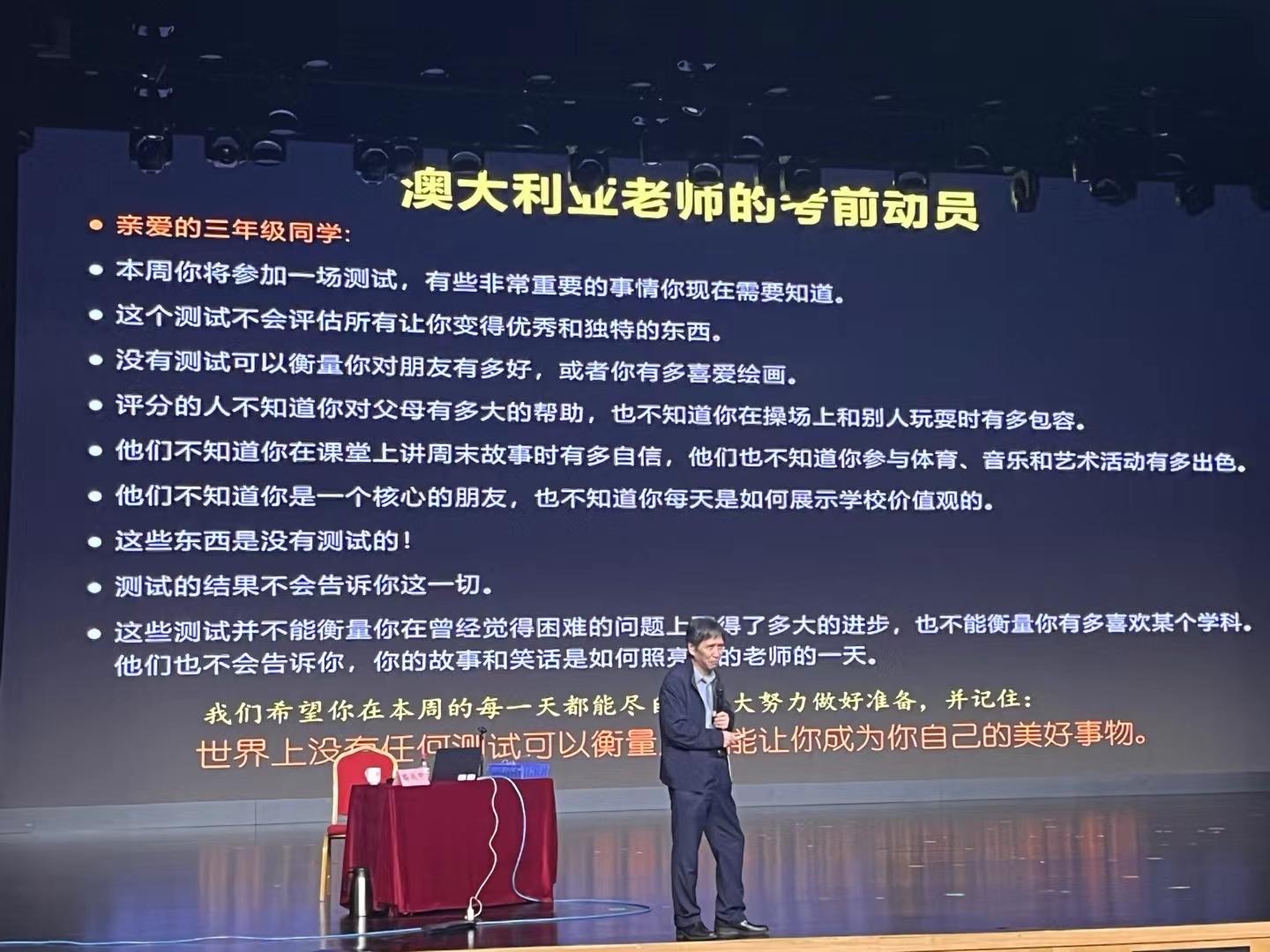 A man in a suit stands on a stage in front of a large screen with Chinese text, possibly giving a presentation or lecture in an auditorium or conference setting. (Captioned by AI)