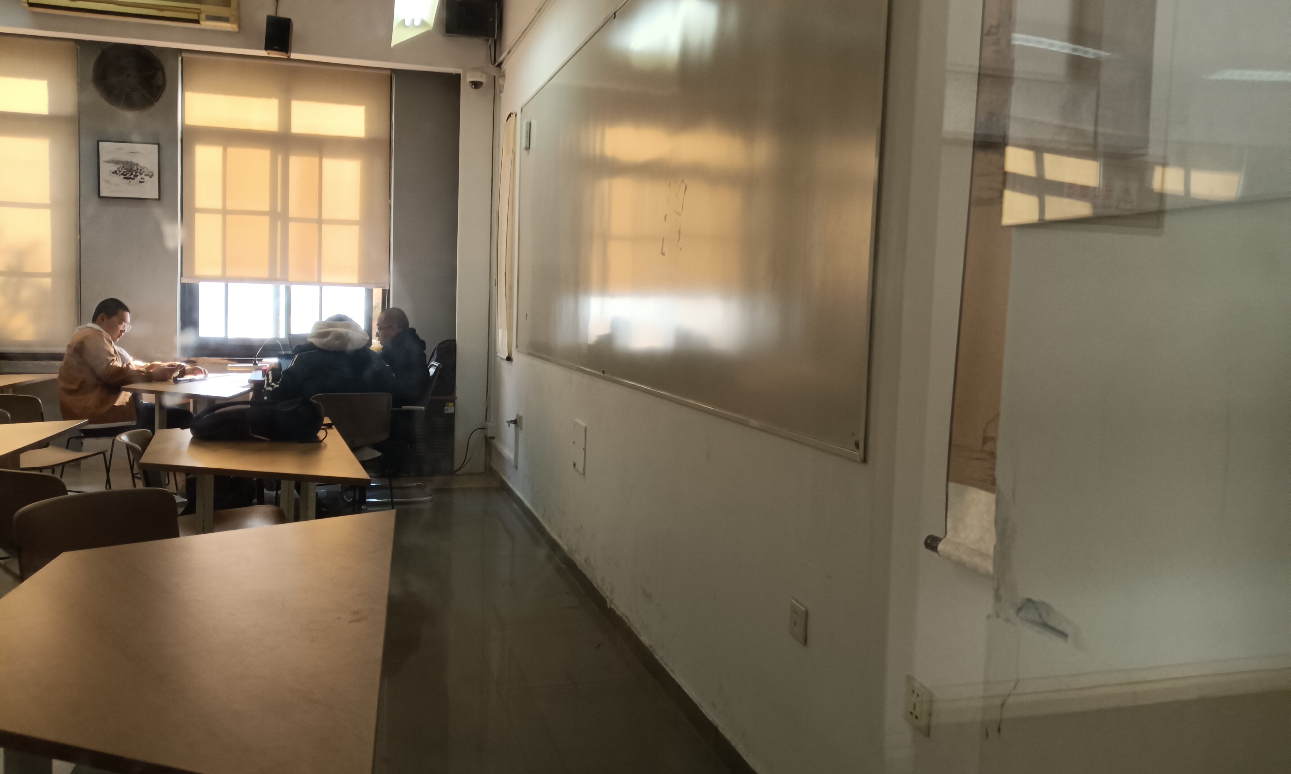 The photo shows a mostly empty classroom with two students working at a desk near a window and a whiteboard on the opposite wall. (Captioned by AI)