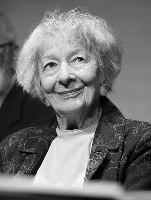 A black and white portrait captures an elderly woman with a kind smile, wearing a light-colored shirt under a patterned jacket. (Captioned by AI)