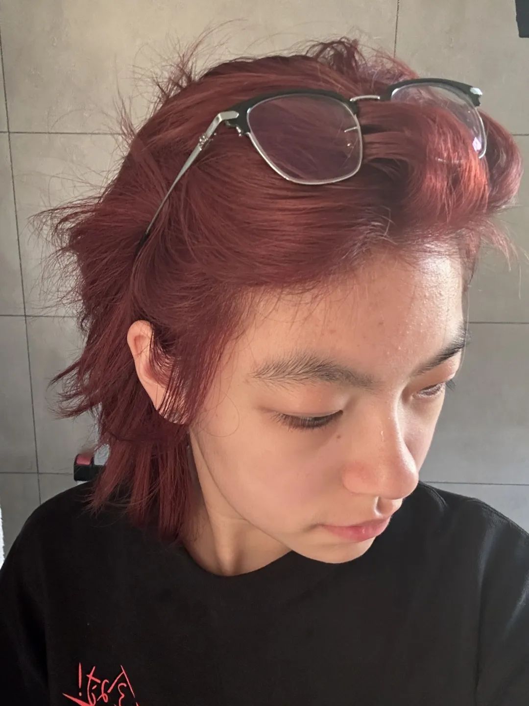 A person with short, auburn hair and glasses perched on their head looks down while wearing a black t-shirt. (Captioned by AI)