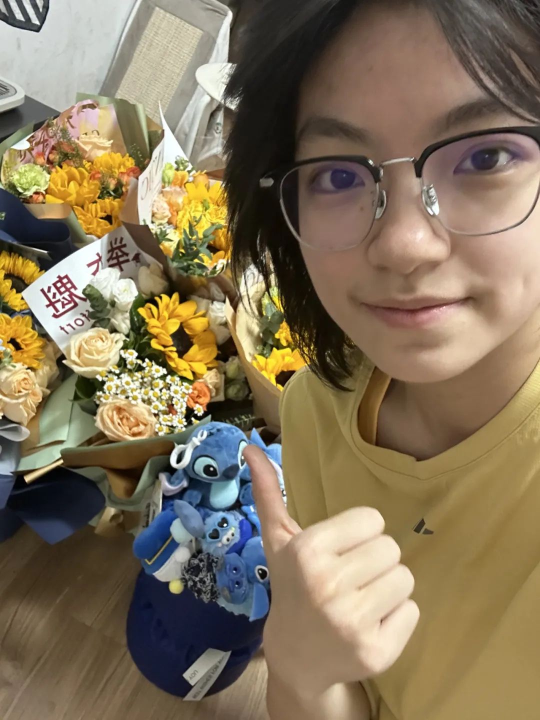 A young person wearing glasses and a yellow shirt gives a thumbs up while surrounded by bouquets of flowers and a Stitch plush toy, possibly celebrating a graduation. (Captioned by AI)