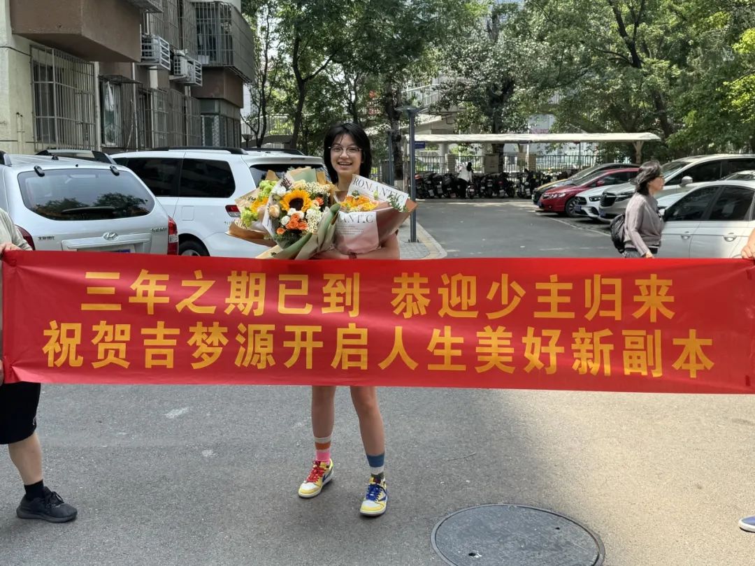 A young woman is being welcomed home with a banner and flowers, possibly after being released from prison after a three-year sentence. (Captioned by AI)